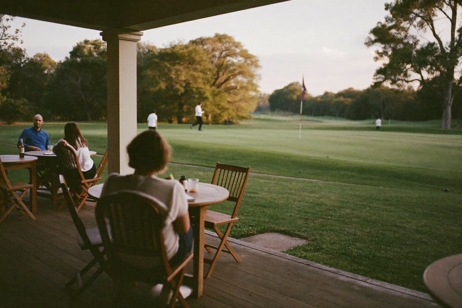 Golfers on a clubhouse terrace looking out over the 18th green in warm evening light